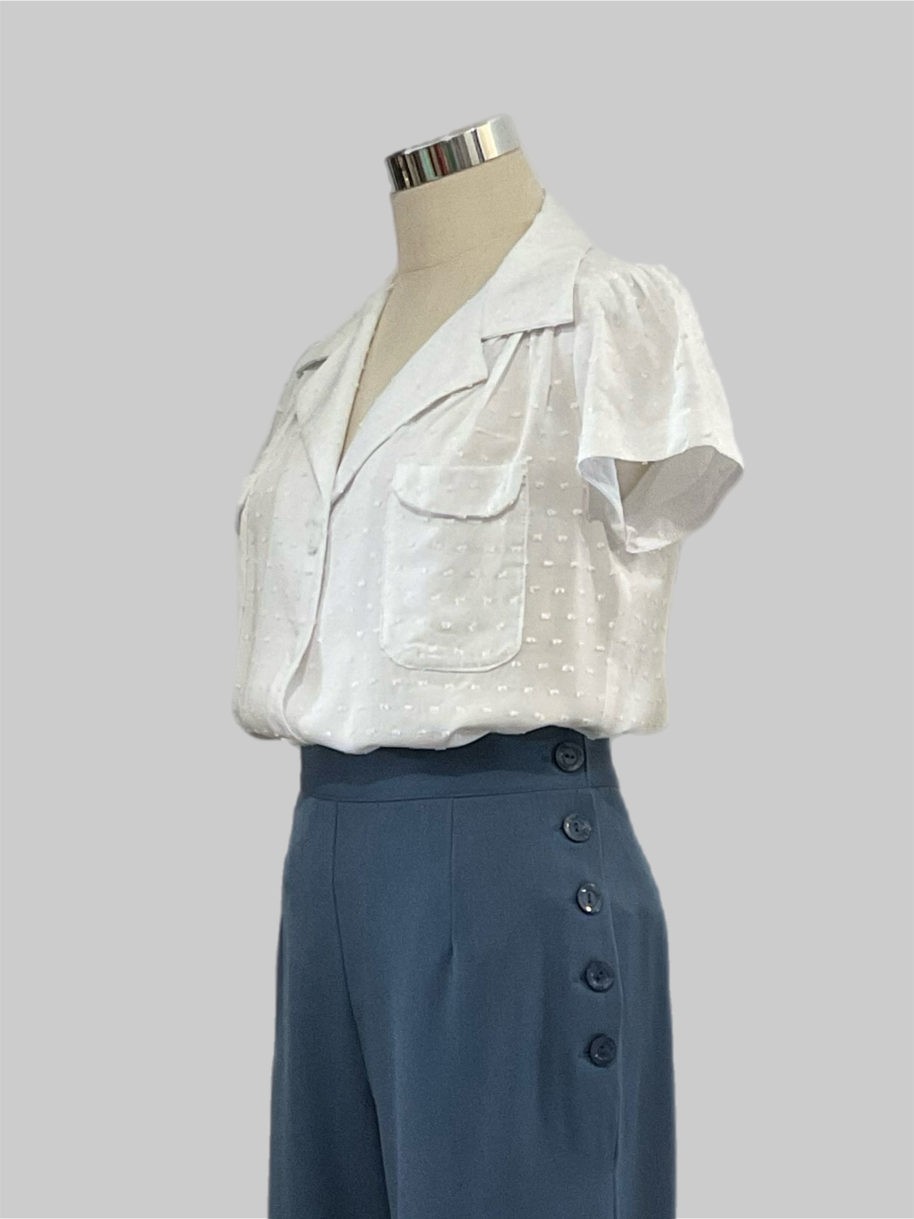 Evita white textured 1940s vintage style blouse with short sleeves and patch pockets displayed on a mannequin wearing high-waisted blue pants.