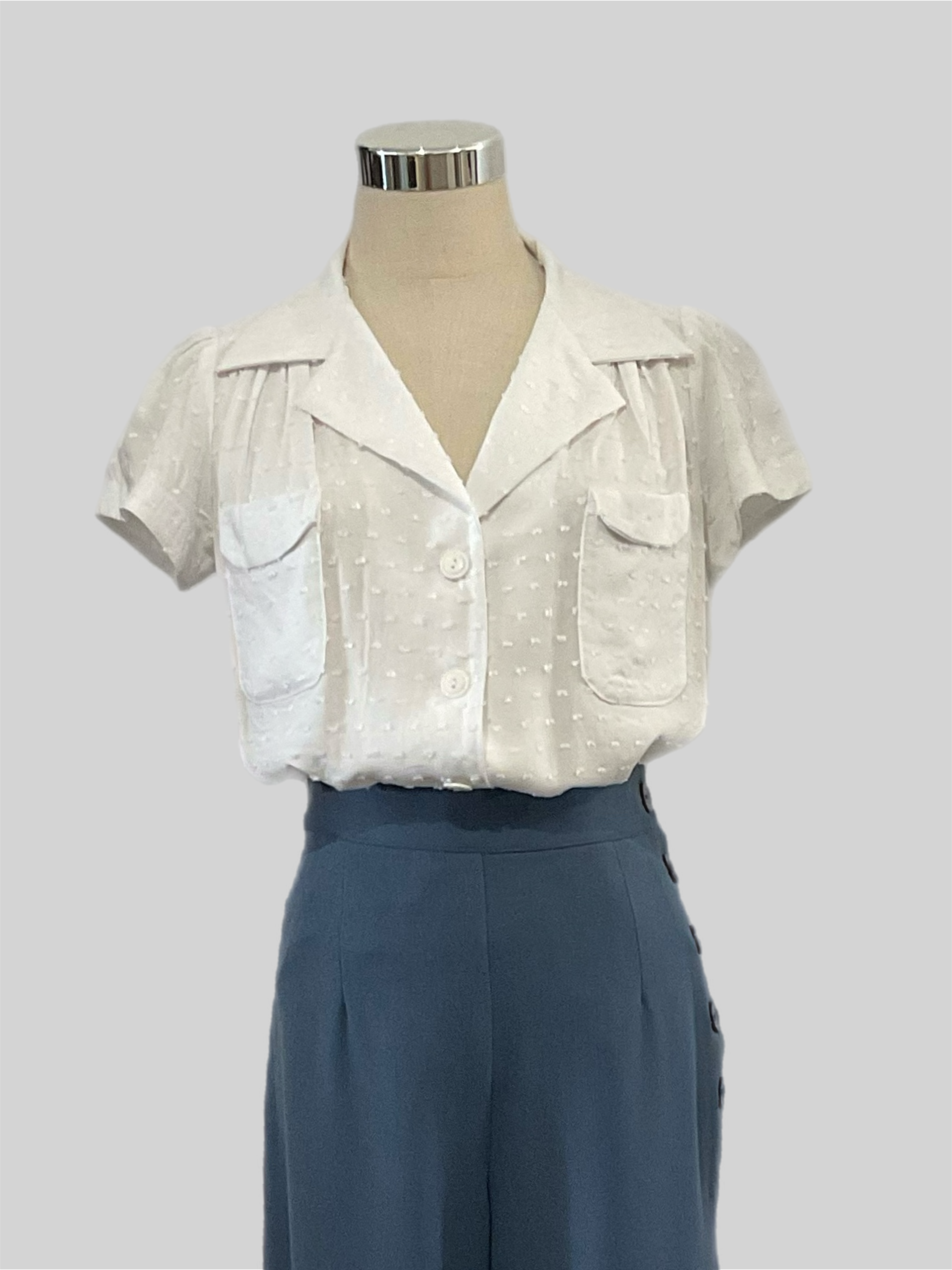 Evita white textured 1940s vintage style blouse with short sleeves and patch pockets displayed on a mannequin wearing high-waisted blue pants.