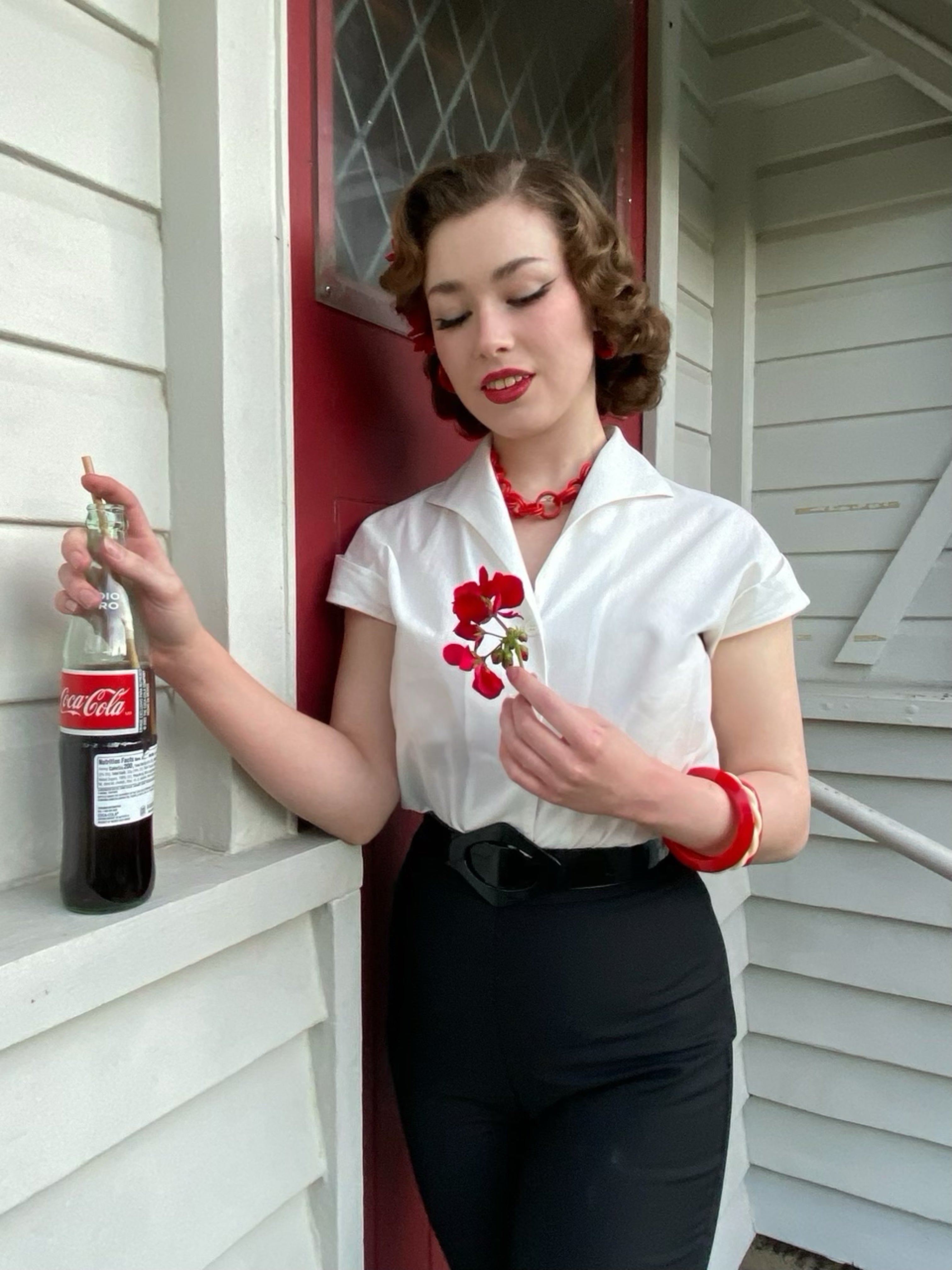 Tammy Natural White Blouse and black Cigarette Pants. Woman in vintage attire holding a Coca-Cola bottle against a house exterior.