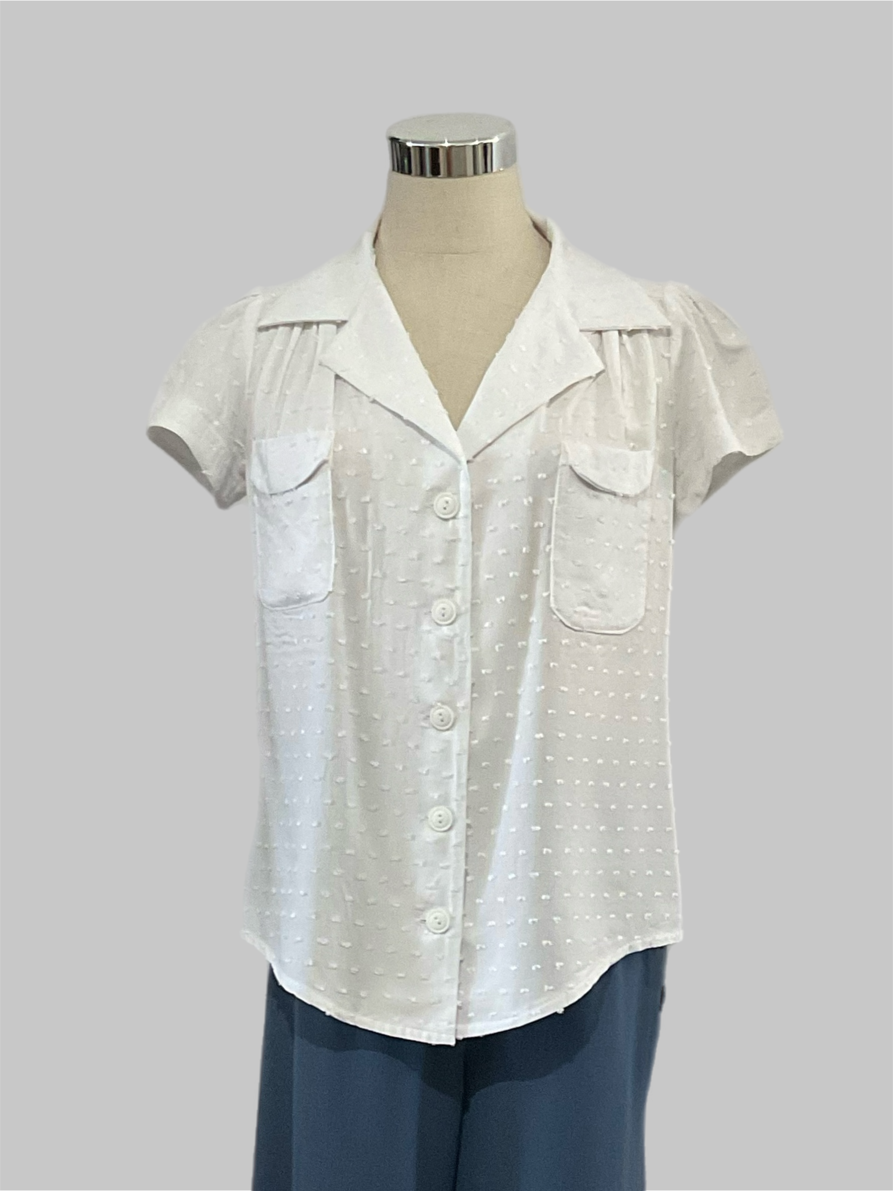 Evita white textured 1940s vintage style blouse with short sleeves and patch pockets displayed on a mannequin wearing high-waisted blue pants.